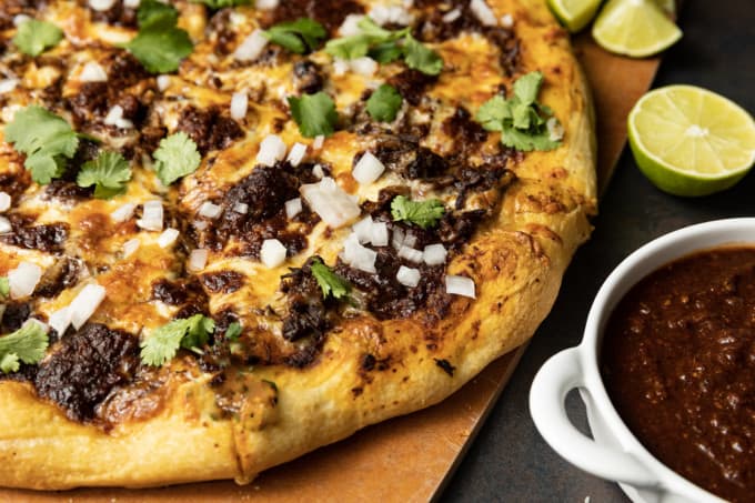 Birria Pizza: Chewy crust topped with tender shredded birria beef, smoky birria stew, loads of melted cheese, chopped sweet onion, fresh cilantro, a squeeze of fresh lime, and a bowl of red chile sauce for dipping. Imagine the best tacos you’ve ever tasted turned into a pizza and you have an idea just how fabulous this pizza is.