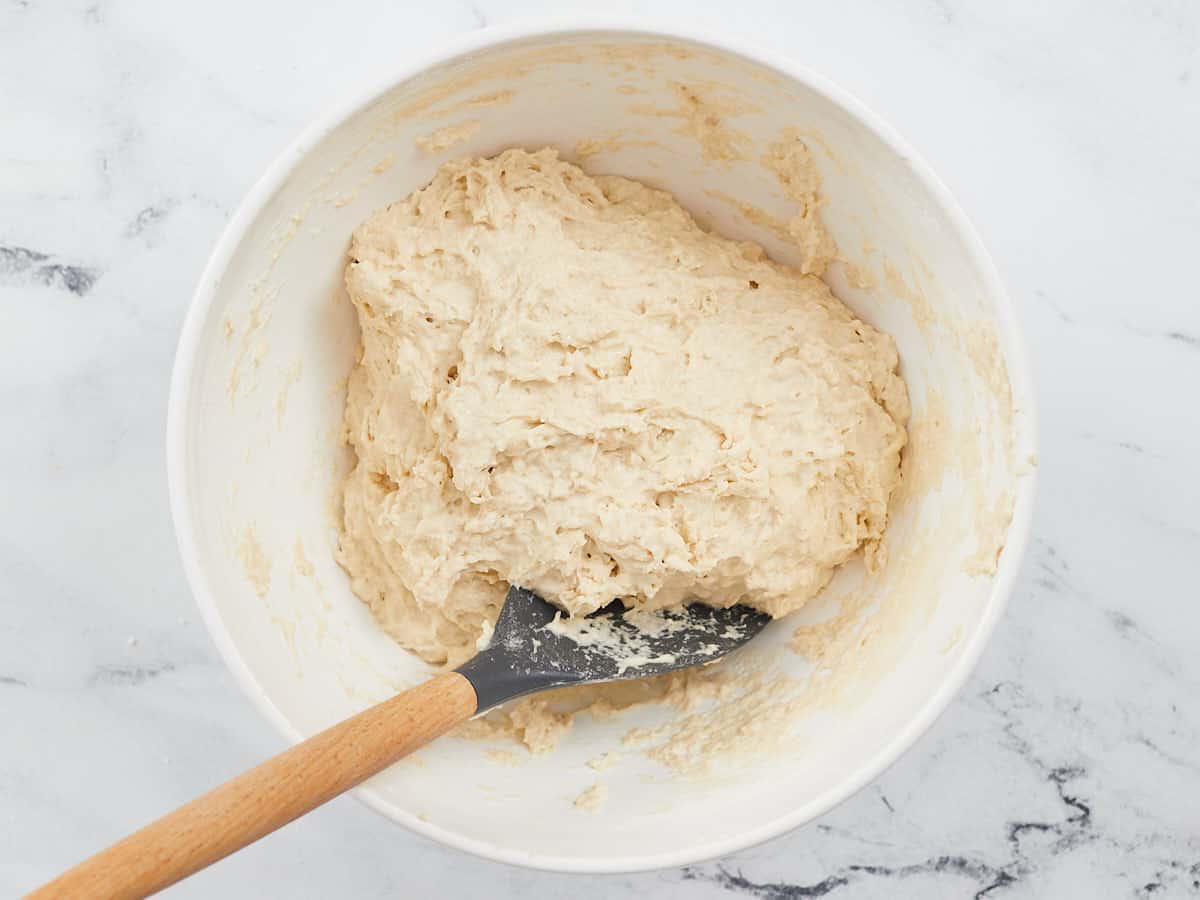 Ingredients for a foccacia-style pizza crust mixed together in a white bowl with a wooden spatula sticking out of the bottom left side of the bowl.