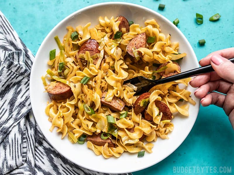 A hand lifting a forkful of Cajun Cabbage and Noodles from a large bowl.