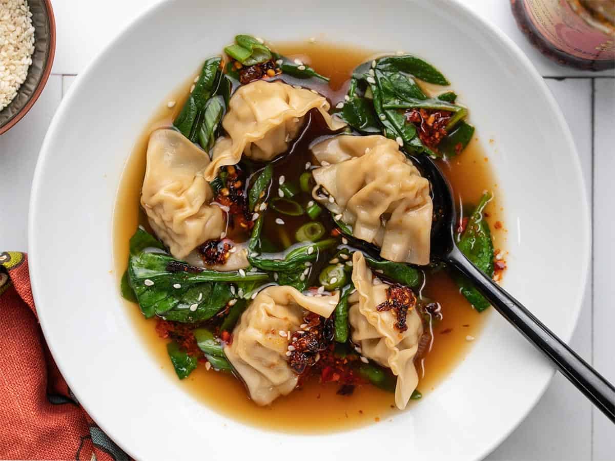 A close up view of dumpling soup in a white bowl topped with white sesame seeds and chili crisp.