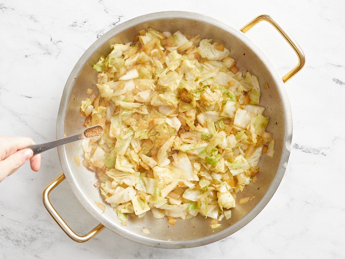 Wilted cabbage in the skillet, seasoning being sprinkled over top. 